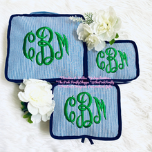 Load image into Gallery viewer, Three matching fabric pouches or small bags with blue-and-white vertical stripes and green embroidered monogram lettering, arranged on a white faux-fur surface with white flowers as props. A pink watermark text is visible across the center.