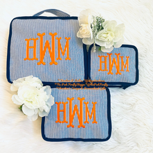 Load image into Gallery viewer, Three matching fabric pouches or small bags with a blue and white striped pattern and orange monogram lettering, arranged on a white faux-fur surface with white flowers as props.
