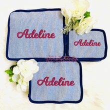 Load image into Gallery viewer, Three personalized fabric pouches with blue and white vertical stripes, navy blue borders, and the name 'Adeline' embroidered in pink on each, arranged on a white textured background with white flowers.
